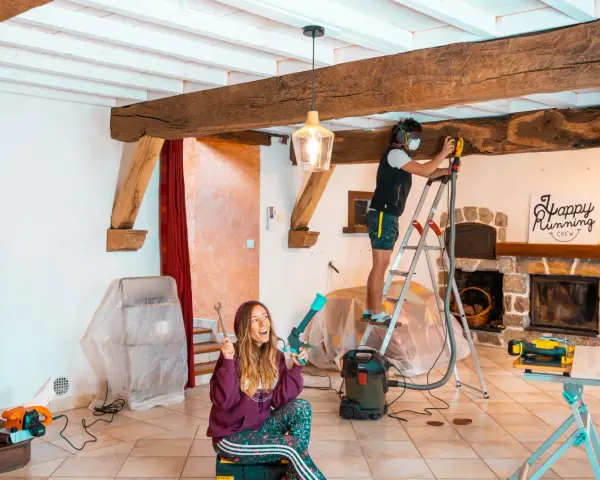 Un homme ponce une poutre en bois avec une ponceuse, tandis qu'une femme tient des outils.