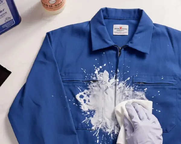 Une main gantée essaie de nettoyer peinture acrylique sèche sur une veste bleue avec un chiffon. Un grattoir et un produit sont à proximité.