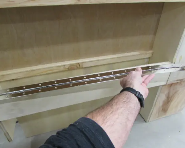 Main tenant une longue charnière piano pour la fixer sur un meuble en bois.