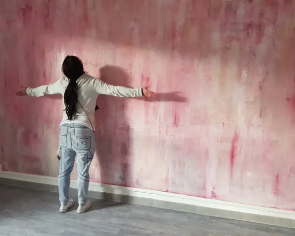 Une personne admire une peinture à la spatule sur mur aux tons roses et blancs, les bras écartés.