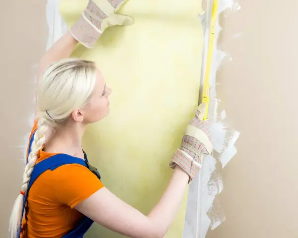 Une femme en salopette bleue et t-shirt orange mesure un mur pour le détapisser puis peindre.