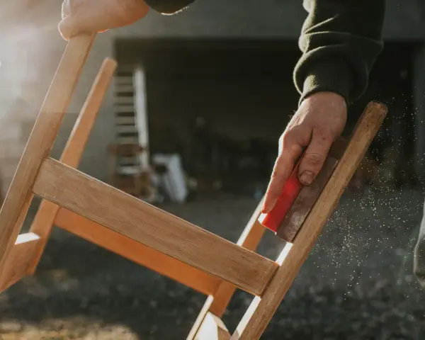 Main d'artisan ponçant un meuble en bois, préparant la surface pour enlever peinture sur bois acétone. Poussière de bois en suspension.