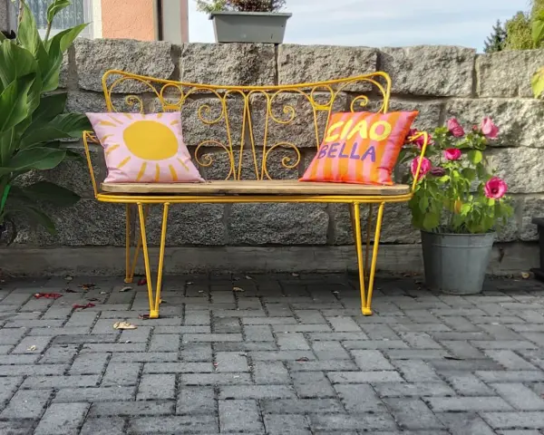 Banc en fer forgé jaune vif, repeint, avec coussins décoratifs. Un soleil rose et "Ciao Bella" sur fond rayé.
