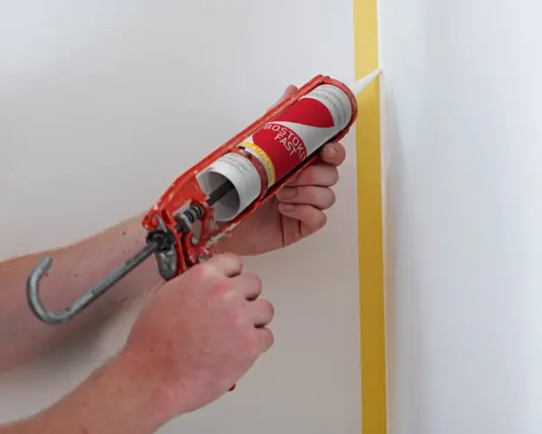 Application de silicone avec un pistolet pour une finition parfaite. Le joint de silicone est appliqué le long d'un coin de mur masqué par du ruban de peintre.