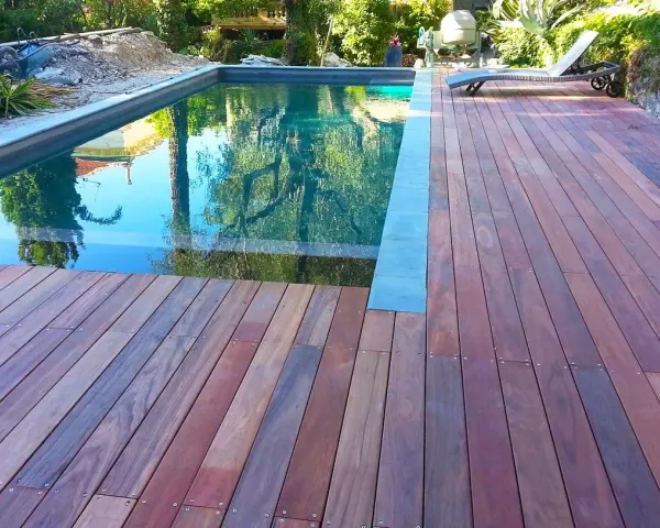 Terrasse en padouk vieilissant, reflétant les arbres dans la piscine. Un transat attend au soleil.