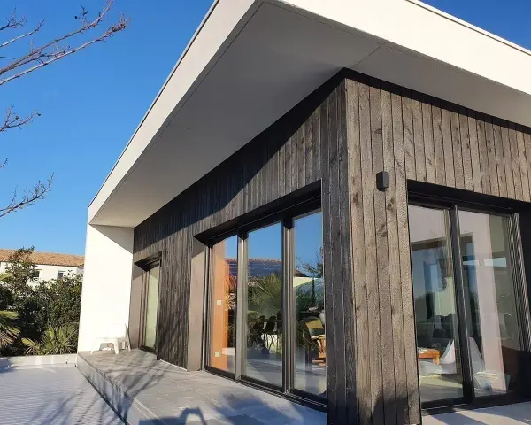 Façade moderne avec bardage en **bois brulé**, grandes baies vitrées reflétant le ciel bleu et la végétation.