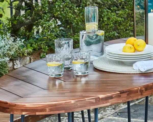 Table en bois couleur acacia, prête pour un apéritif en plein air avec des verres d'eau citronnée, des citrons et une bougie.