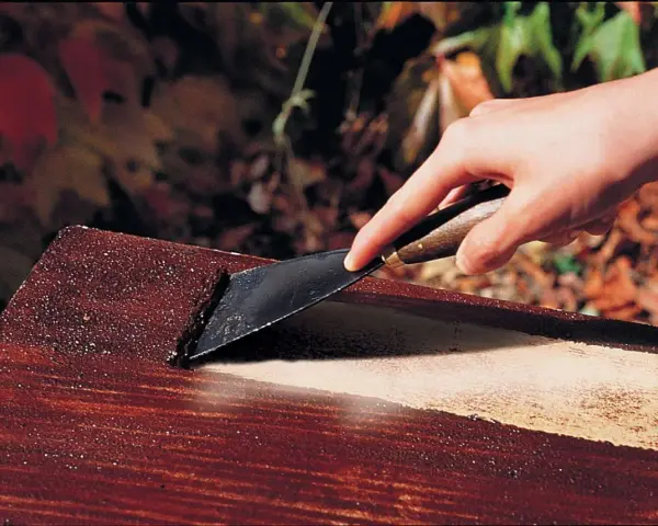 Main d'une personne utilisant un grattoir pour enlever de la peinture d'un meuble en bois, comme avec un décapant peinture bois fait maison.