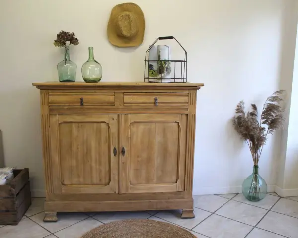 Buffet en bois clair, prêt pour sa restauration. Deux tiroirs et deux portes ornées de moulures. Décoration champêtre avec des vases anciens et des herbes sèches.