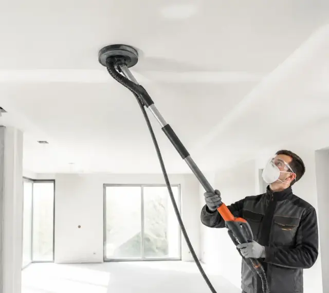 Un ouvrier utilise une ponceuse girafe pour poncer un plafond blanc dans une pièce en construction.