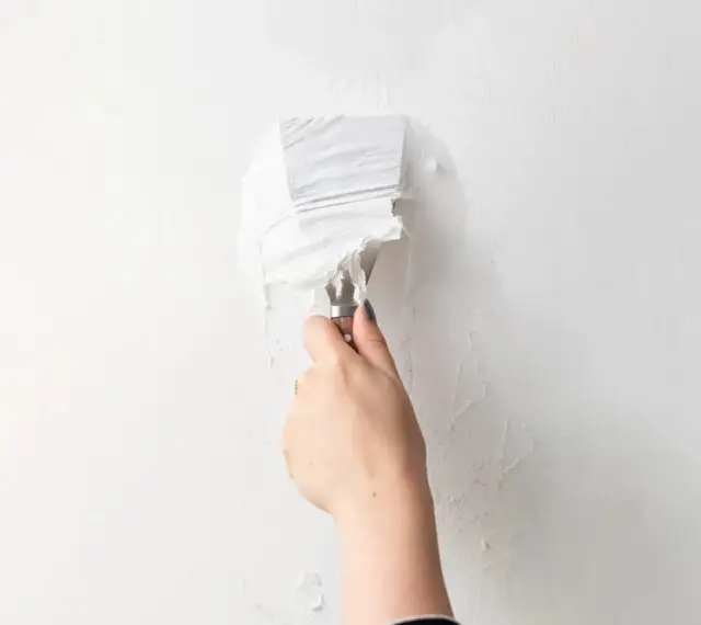 Une main utilise un couteau à mastic pour enlever de la vieille peinture écaillée d'un mur blanc, une étape essentielle pour savoir comment enlever la colle sur un mur avant de peindre.