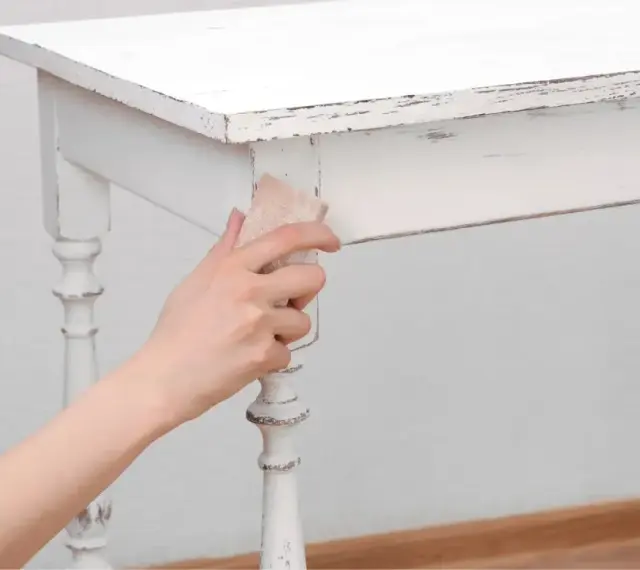 Main tenant enlever peinture sur bois d'une table blanche vieillie, révélant le grain du bois.