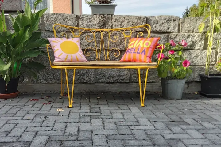 Banc en fer forgé jaune vif, repeint, avec coussins décoratifs. Un soleil rose et "Ciao Bella" sur fond rayé.