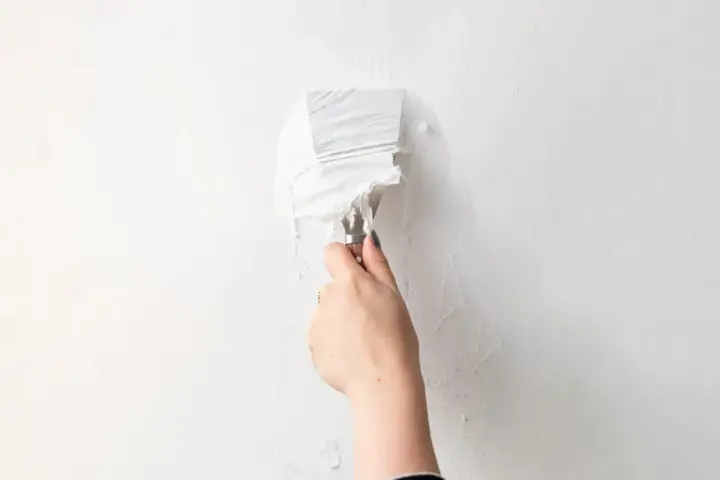 Une main utilise un couteau à mastic pour enlever de la vieille peinture écaillée d'un mur blanc, une étape essentielle pour savoir comment enlever la colle sur un mur avant de peindre.