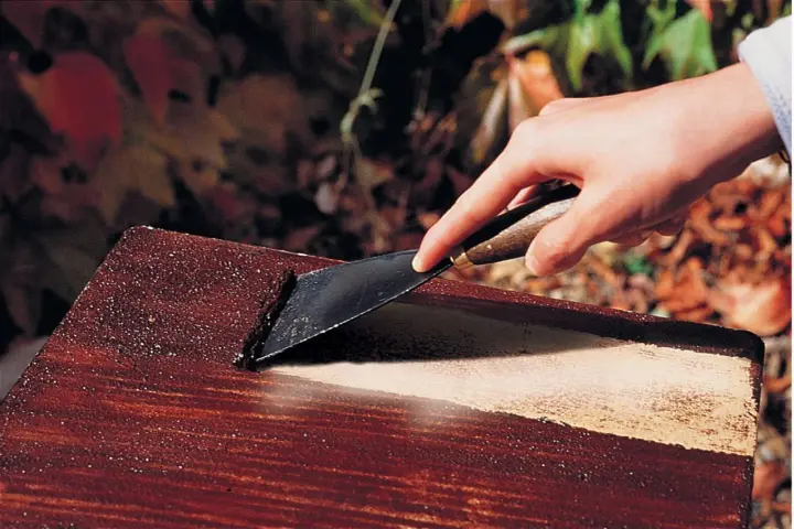 Main d'une personne utilisant un grattoir pour enlever de la peinture d'un meuble en bois, comme avec un décapant peinture bois fait maison.