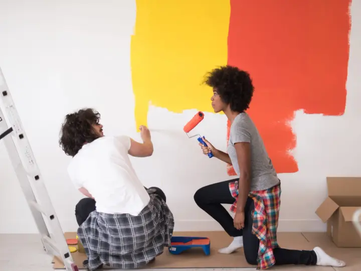 Un couple peint un mur en jaune et orange. Ils utilisent des rouleaux pour nettoyer peinture glycero. Une échelle est à gauche.