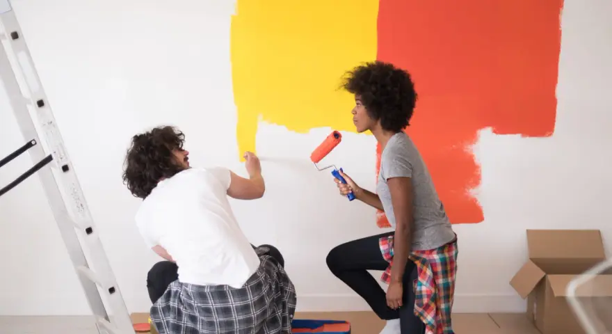 Un couple peint un mur en jaune et orange. Ils utilisent des rouleaux pour nettoyer peinture glycero. Une échelle est à gauche.