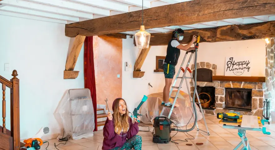 Un homme ponce une poutre en bois avec une ponceuse, tandis qu'une femme tient des outils.