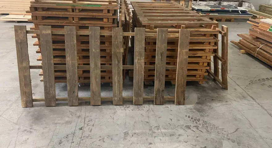 Des planches de bois recycle, issues de palettes, forment une barrière devant d'autres palettes empilées.