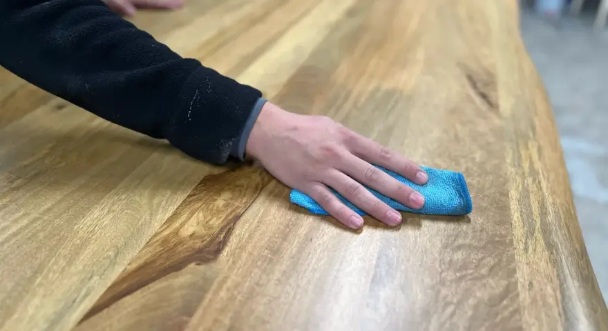 Main d'une personne passant un chiffon bleu sur une table en bois, montrant comment nettoyer une table en bois.