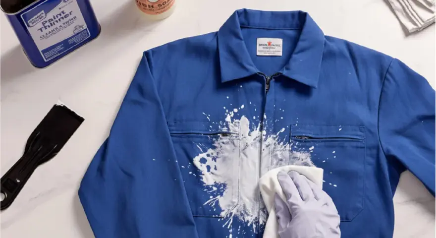 Une main gantée essaie de nettoyer peinture acrylique sèche sur une veste bleue avec un chiffon. Un grattoir et un produit sont à proximité.