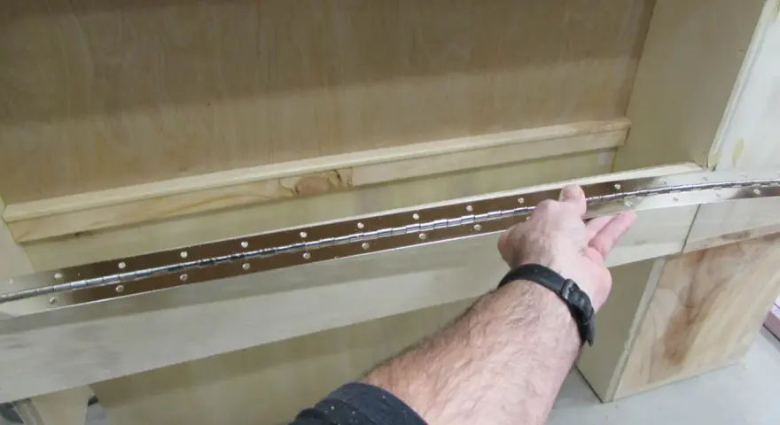 Main tenant une longue charnière piano pour la fixer sur un meuble en bois.