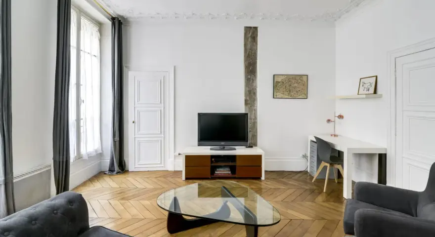 Salon moderne avec parquet en chevrons, canapés gris, bureau et TV. Les murs et le plafond sont peints à l'huile, offrant une finition impeccable.