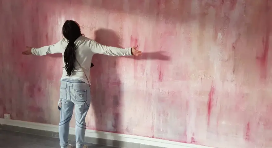 Une personne admire une peinture à la spatule sur mur aux tons roses et blancs, les bras écartés.