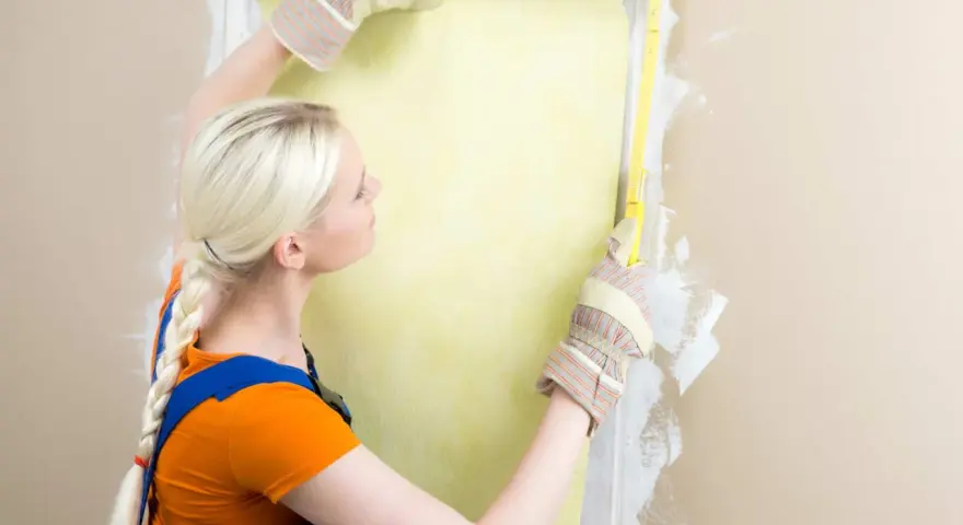 Une femme en salopette bleue et t-shirt orange mesure un mur pour le détapisser puis peindre.