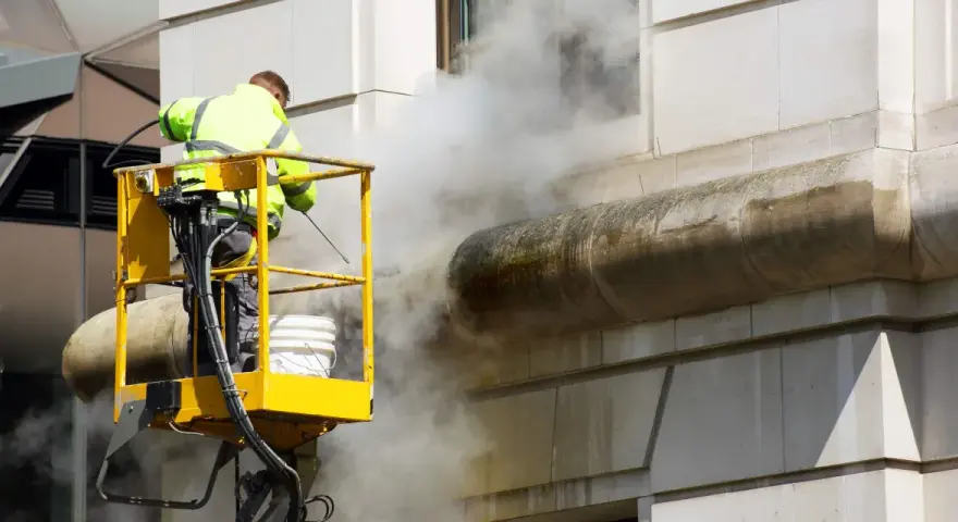 Un ouvrier en nacelle jaune utilise un nettoyeur haute pression pour décaper la peinture d'une façade en pierre, créant un nuage de vapeur.