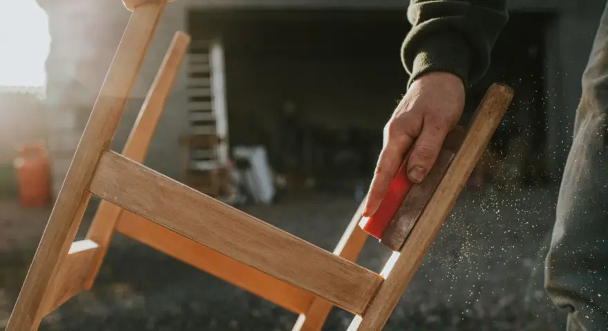 Main d'artisan ponçant un meuble en bois, préparant la surface pour enlever peinture sur bois acétone. Poussière de bois en suspension.