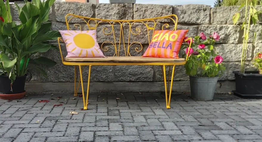 Banc en fer forgé jaune vif, repeint, avec coussins décoratifs. Un soleil rose et "Ciao Bella" sur fond rayé.