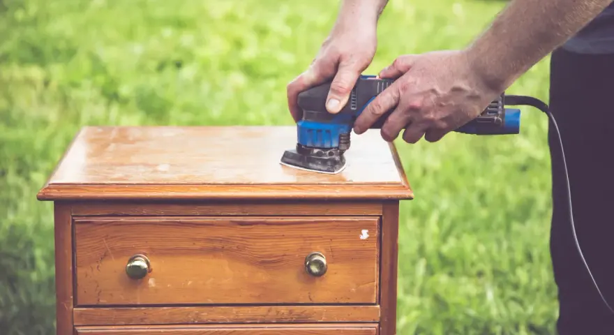 Un homme utilise une ponceuse pour le décapage mécanique d'une commode en bois, préparant le meuble pour une nouvelle finition.