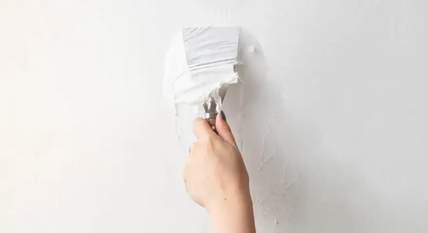 Une main utilise un couteau à mastic pour enlever de la vieille peinture écaillée d'un mur blanc, une étape essentielle pour savoir comment enlever la colle sur un mur avant de peindre.
