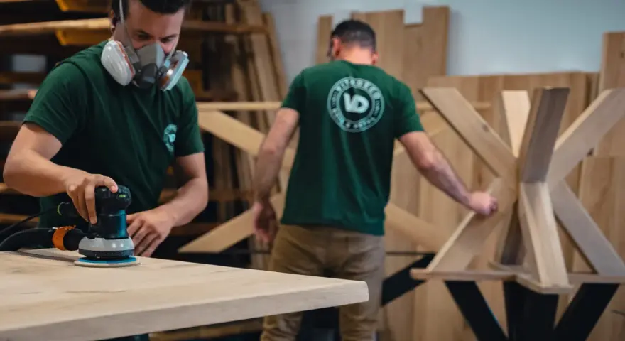 Un artisan, masqué, utilise une ponceuse pour poncer une table en bois. Un autre artisan travaille sur un pied de table en arrière-plan.