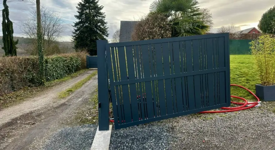 Portail en pente avec gonds sur mesure. Le portail gris foncé est ouvert, révélant un chemin de gravier et une allée bordée de haies.