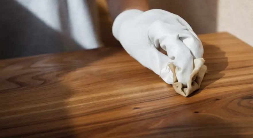 Main d'une personne portant un gant blanc, appliquant de la cire sur un meuble en bois. Apprendre comment cirer un meuble pour le protéger.
