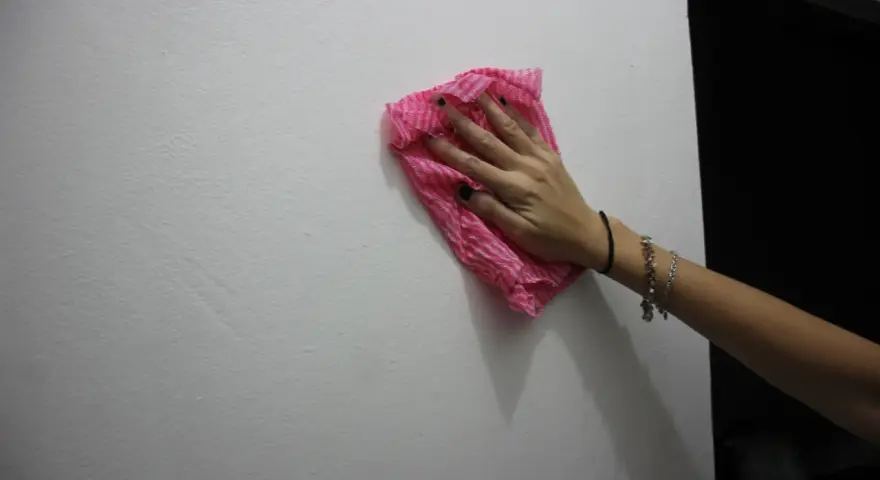 Main tenant une lingette rose pour nettoyer un mur. Une astuce pour savoir comment enlever de la colle forte sur un mur.
