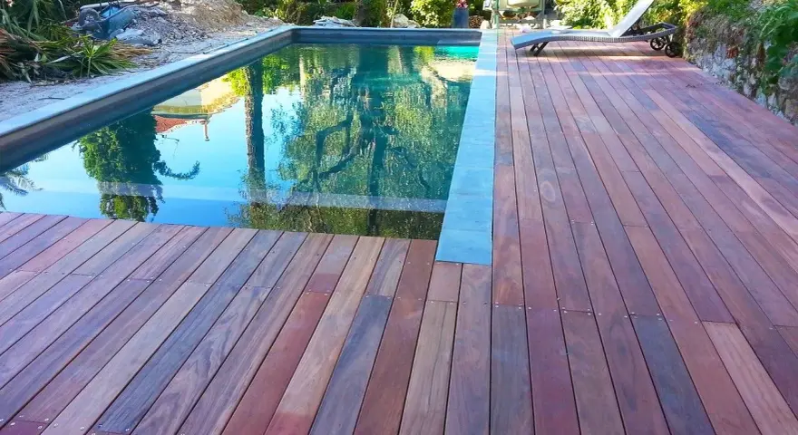 Terrasse en padouk vieilissant, reflétant les arbres dans la piscine. Un transat attend au soleil.