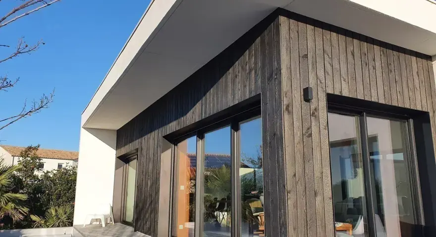 Façade moderne avec bardage en **bois brulé**, grandes baies vitrées reflétant le ciel bleu et la végétation.