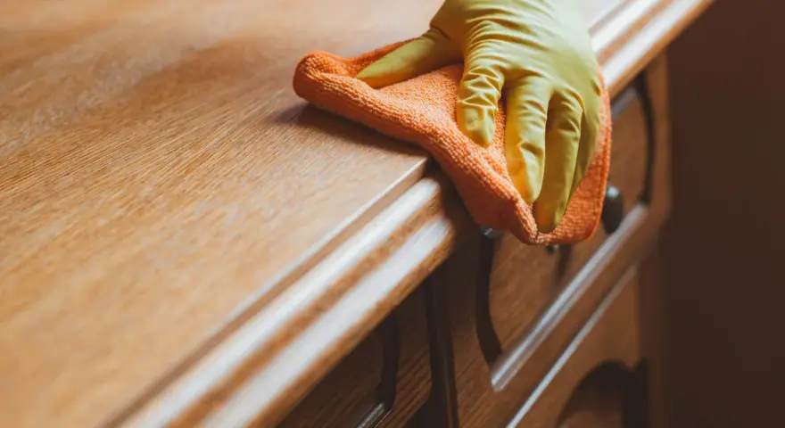 Main d'une personne portant un gant jaune, essuyant un meuble en bois avec un chiffon orange. La qualité du bois est mise en valeur par la lumière.