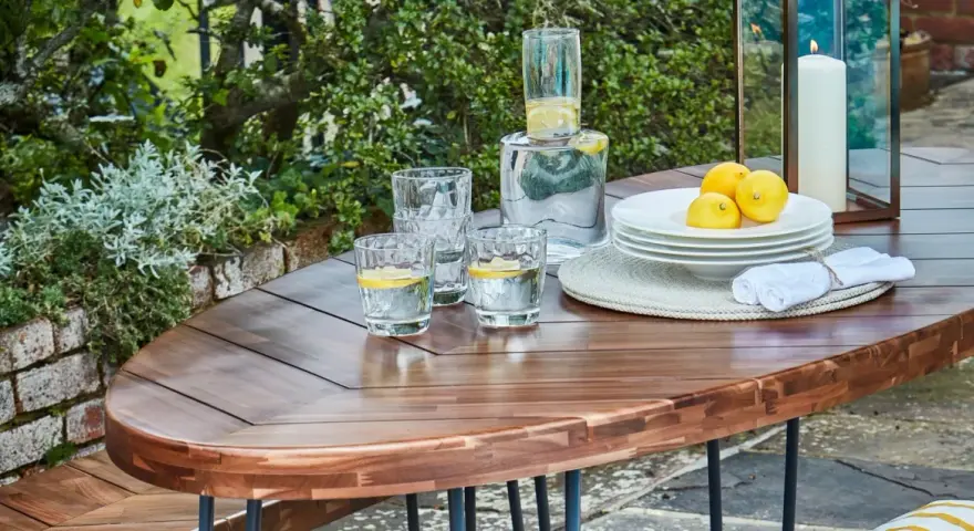 Table en bois couleur acacia, prête pour un apéritif en plein air avec des verres d'eau citronnée, des citrons et une bougie.