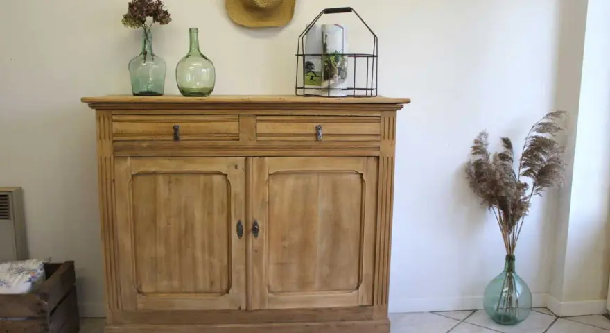 Buffet en bois clair, prêt pour sa restauration. Deux tiroirs et deux portes ornées de moulures. Décoration champêtre avec des vases anciens et des herbes sèches.