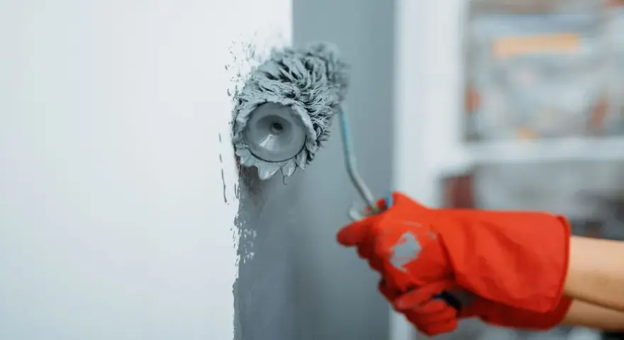 Main tenant d'un rouleau à peinture gris, une main gantée de rouge applique la peinture sur un mur blanc. La technique peinture mur est en cours.