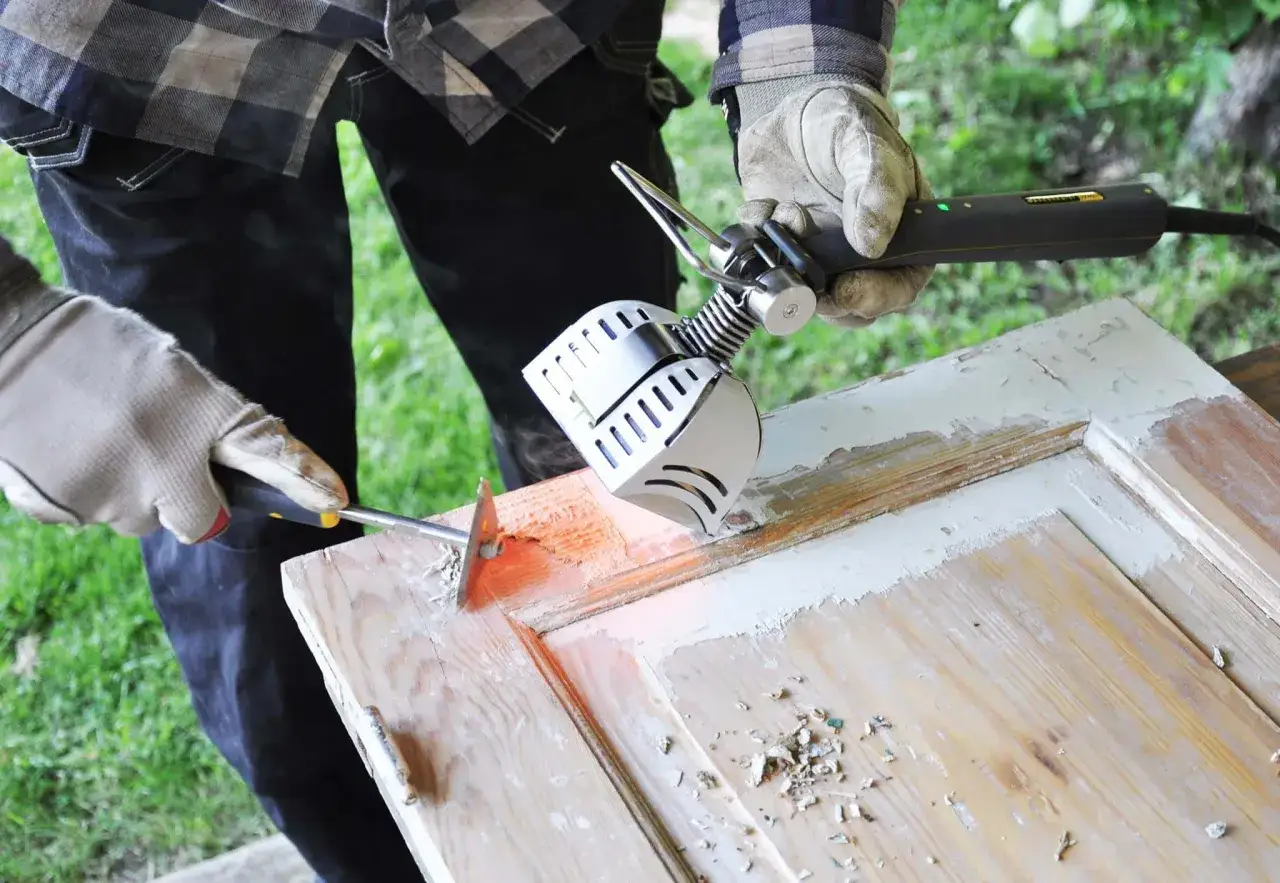 Main tenant enlever peinture sur bois avec un décapeur thermique et une spatule. Des éclats de peinture tombent sur la surface.