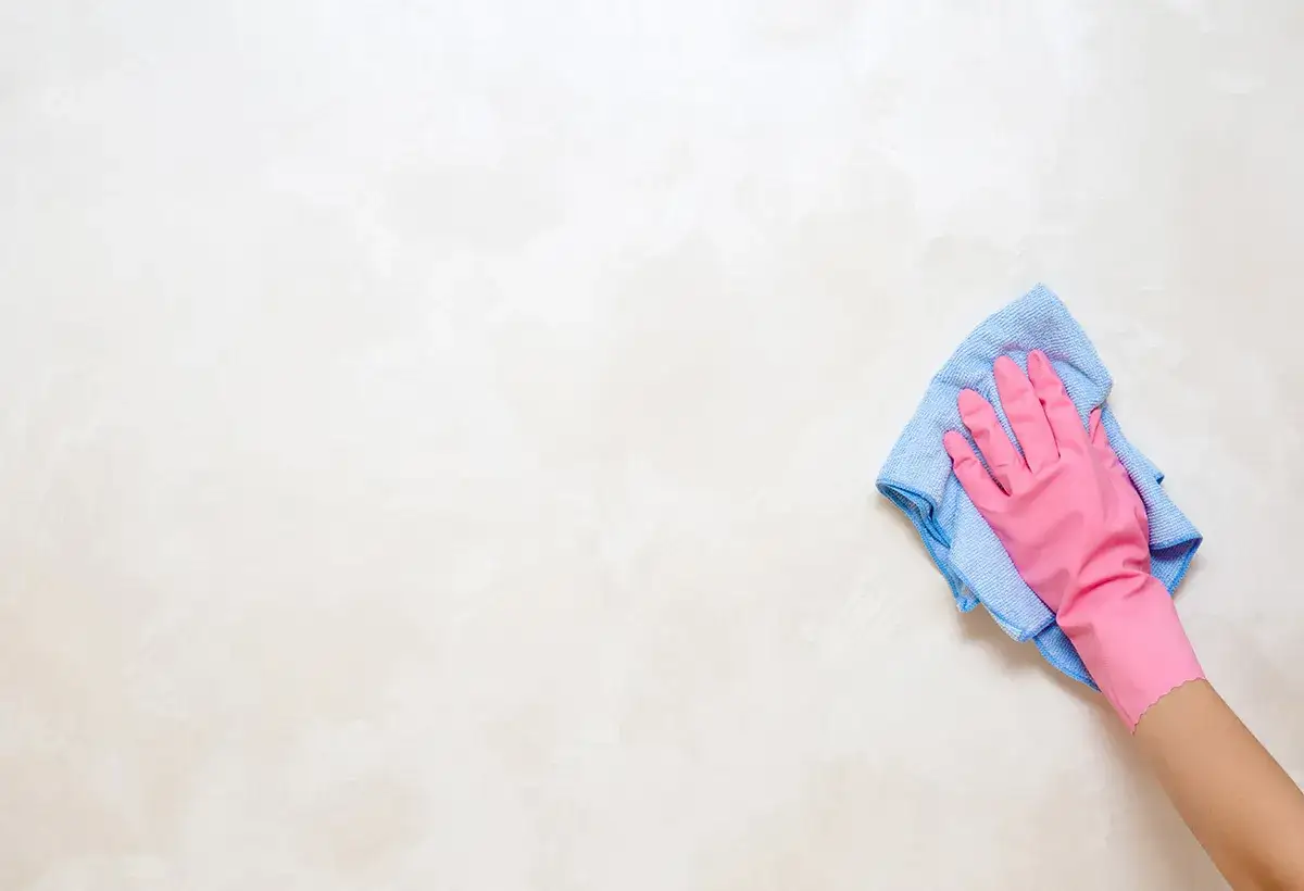 Main rose, nettoyant un mur avec un chiffon bleu. Une étape essentielle pour savoir comment enlever la colle sur un mur avant de peindre.