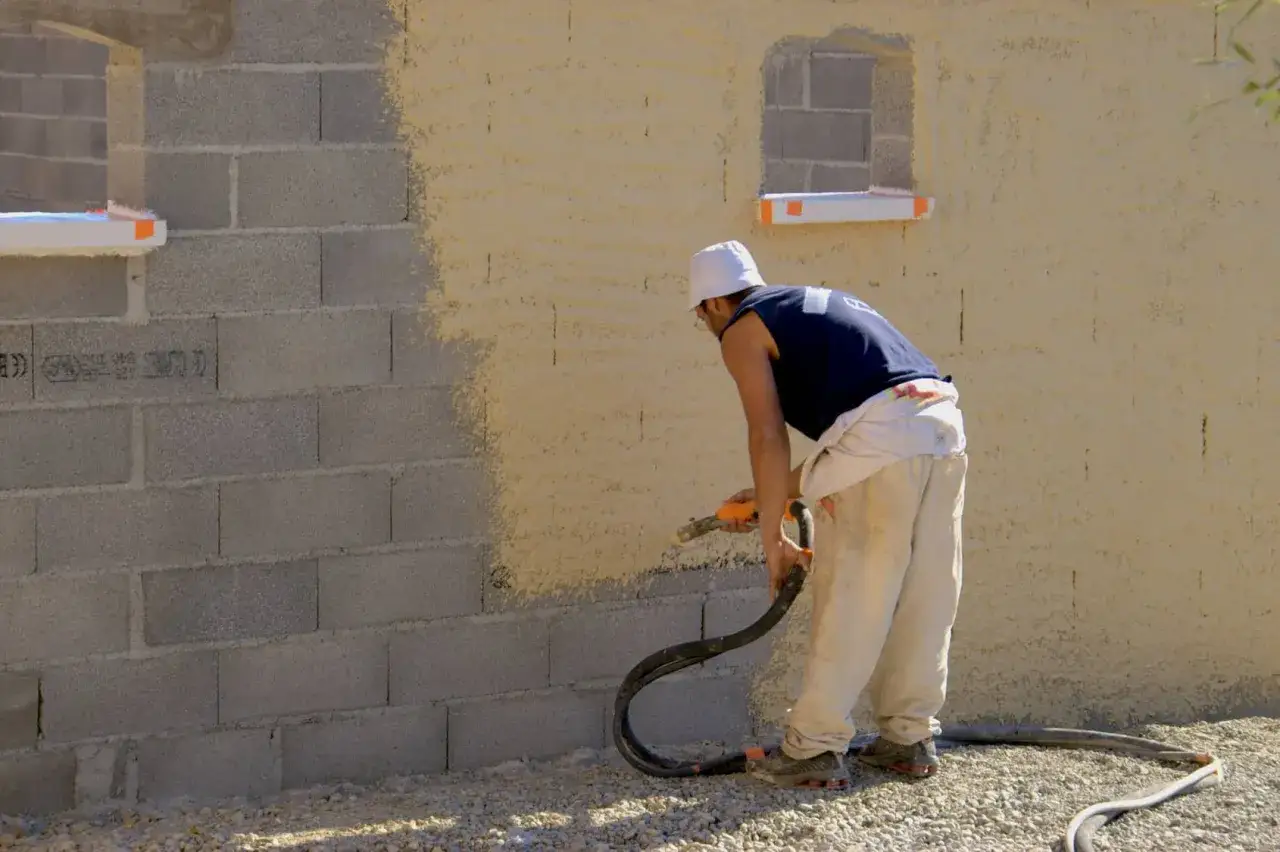 Un ouvrier utilise un pistolet pour peindre un mur en parpaings. La moitié du mur est déjà recouverte d'un enduit texturé de couleur ocre.