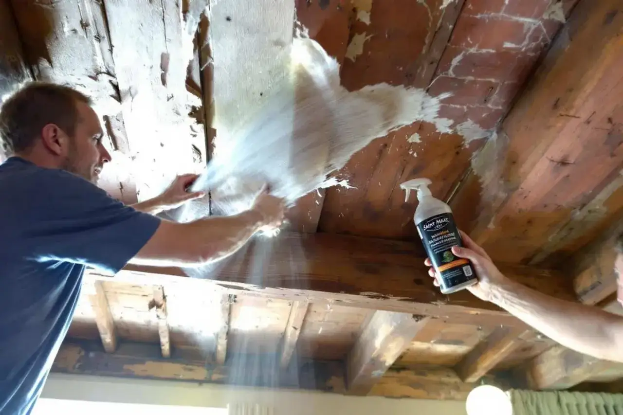 Un homme utilise un produit Saint Marc pour nettoyer des poutres en bois. Le produit semble efficace pour enlever une vieille couche.