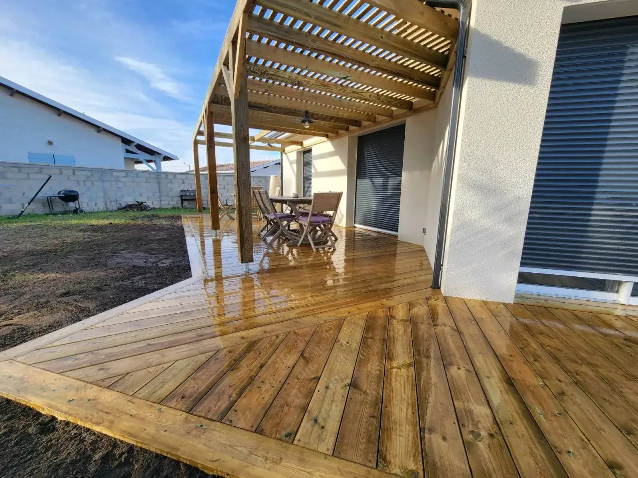 Terrasse en bois classe 3 fra&icirc;chement trait&eacute;e, refl&eacute;tant le ciel bleu. Une table et des chaises attendent sous la pergola.