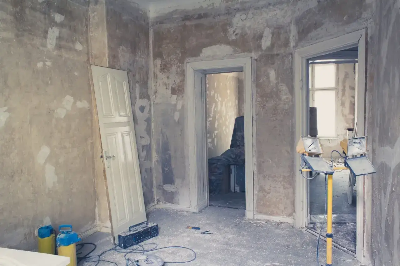 Une pi&egrave;ce en r&eacute;novation, murs en ciment brut, une porte blanche appuy&eacute;e, des projecteurs et une radio sur le sol.